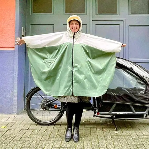 Frau vor einem Lastenrad Arme ausgebreitet mit einem Knielangen Regenponcho für Damen in Grün, vorne mit Reißverschluss, lächelnd.